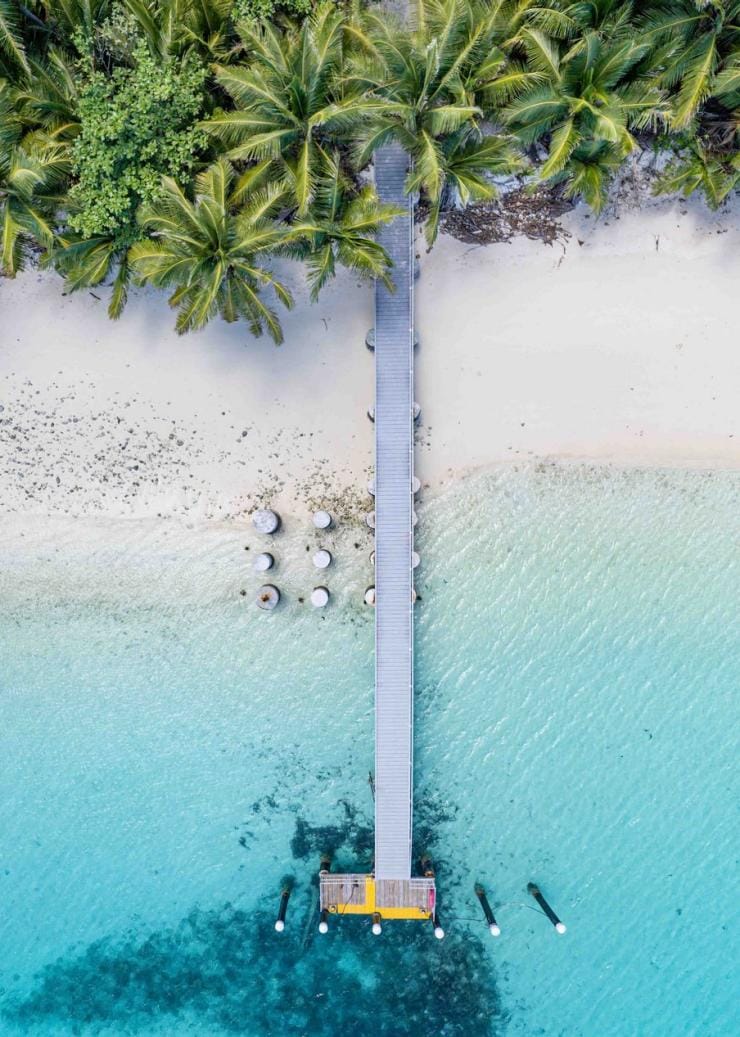 Aerial view over a long, grey pier beginning in the shade of green palm trees before stretching out over a white sand strip and into bright blue water at Cossies Beach, Direction Island, Cocos (Keeling) Islands © Strick & Fran
