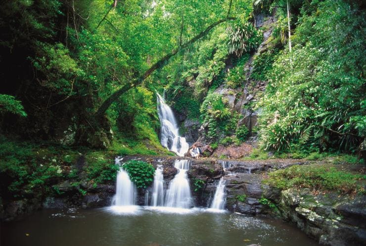 A person crouched down between two cascading waterfalls above and below them while surrounded by greenery at Elebana Falls, Lamington National Park, Queensland © Tourism and Events Queensland