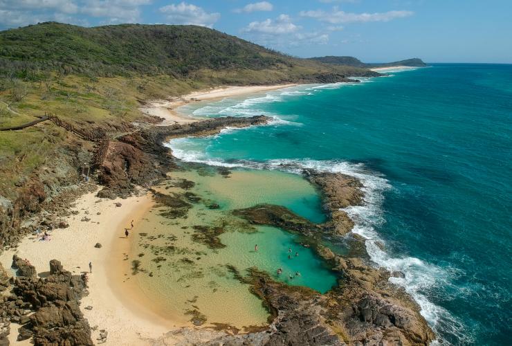 Aerial view over people swimming in the natural formation of the Champagne Pools with waves rolling into the rocks dividing the pools and the ocean on K’gari, Queensland © Tourism Australia