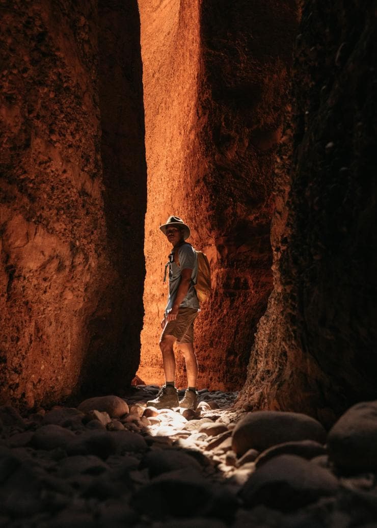 A person standing in the shadows of two red rock walls with a stream of light peaking throw in Echidna Chasm, Purnululu National Park, Western Australia © Tourism Western Australia