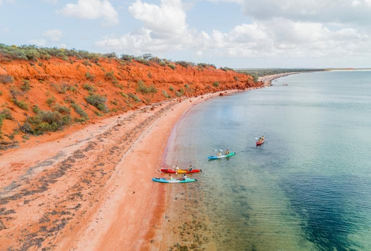 People kayaking on clear blue waters towards a red sandy shore and rock face with Wula Gura Nyinda Eco Adventures, Shark Bay World Heritage Area, Western Australia © Tourism Western Australia