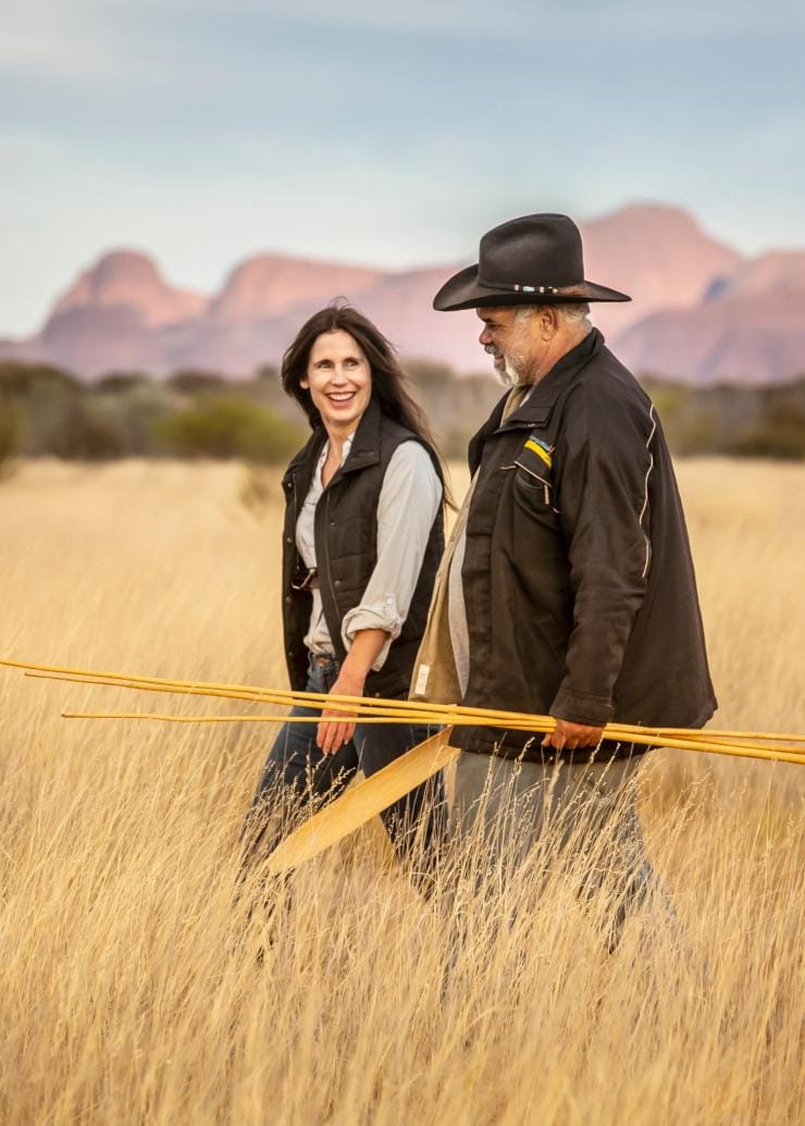 A woman walking through a grassy plain alongside a local Aboriginal guide who is holding artefacts during a cultural tour with SEIT Outback Australia, Uluru, Northern Territory © Tourism NT/Archie Sartracom