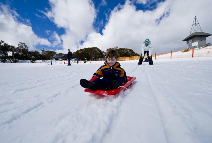 A mother watching from atop a snowy slope as her child rides downhill on a sled while smiling at Mt Buller, Victoria © Visit Victoria