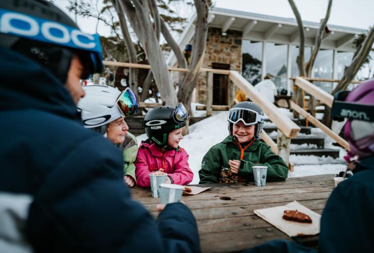 A family smiling over cookies and hot chocolate at a picnic table in the snow, Mt Hotham, High Country, Victoria © Tourism North East