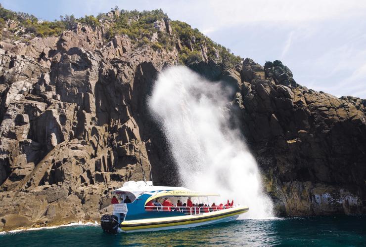 A boat filled with people resting on the ocean beneath rugged, rocky cliffs with a giant spurt of water erupting in front of them during a tour with Bruny Island Cruises, Breathing Rock, Bruny Island, Tasmania © Tourism Tasmania/Glen Gibson