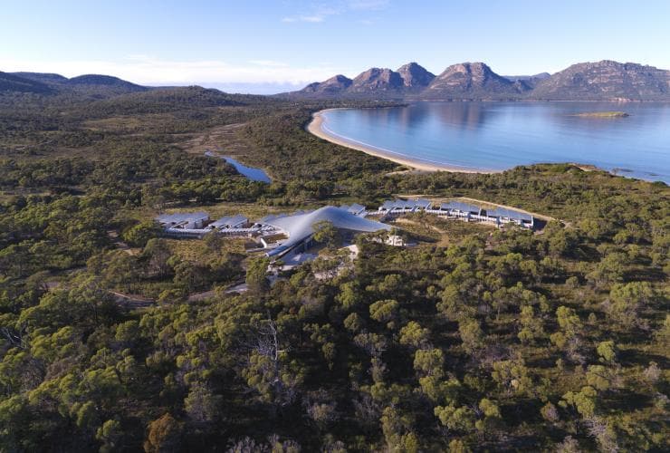 Aerial view over a curved row of buildings surrounded by trees, mountains and the ocean at Saffire Freycinet, Freycinet, Tasmania © Saffire Freycinet