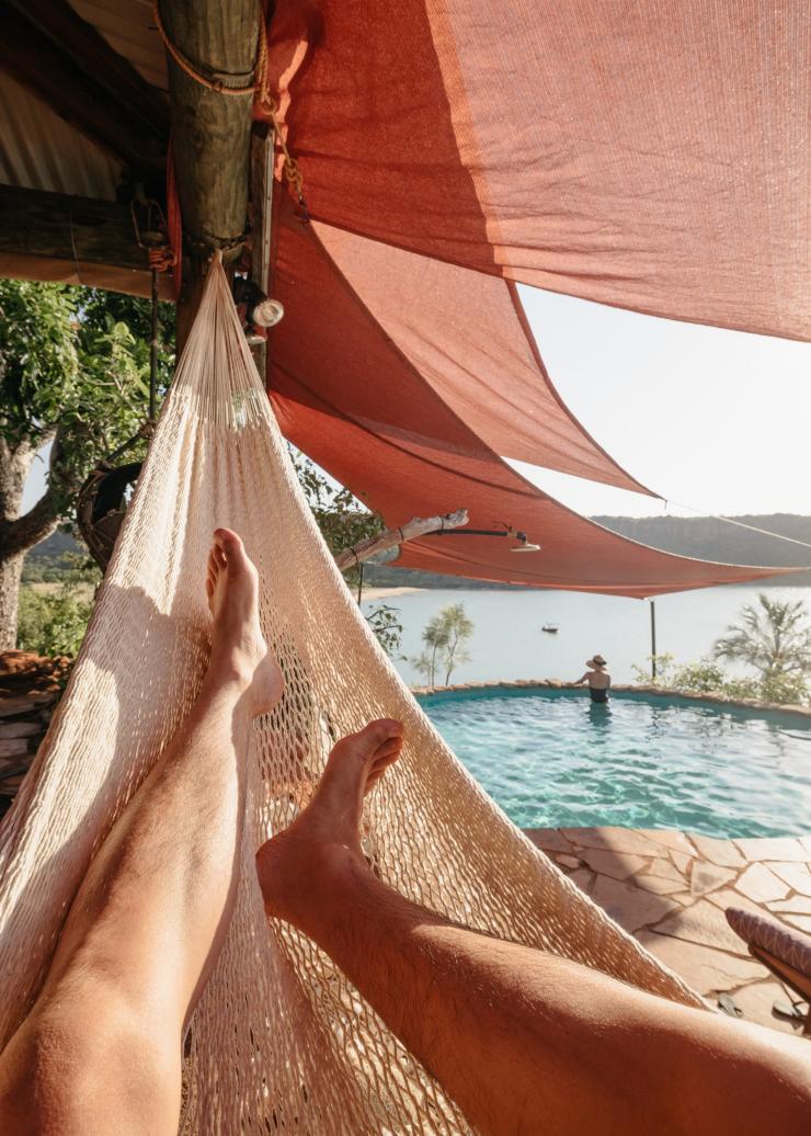 A person relaxing in a hammock in the foreground as another person swims in a pool overlooking the ocean at Faraway Bay, the Kimberley, Western Australia © Tourism Western Australia