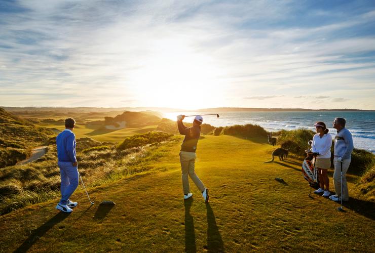 A group of people watching as a man hits a golf ball with kangaroos nearby and the ocean in the background at Barnbougle Lost Farm Golf Links, Bridport, Tasmania © Tourism Australia