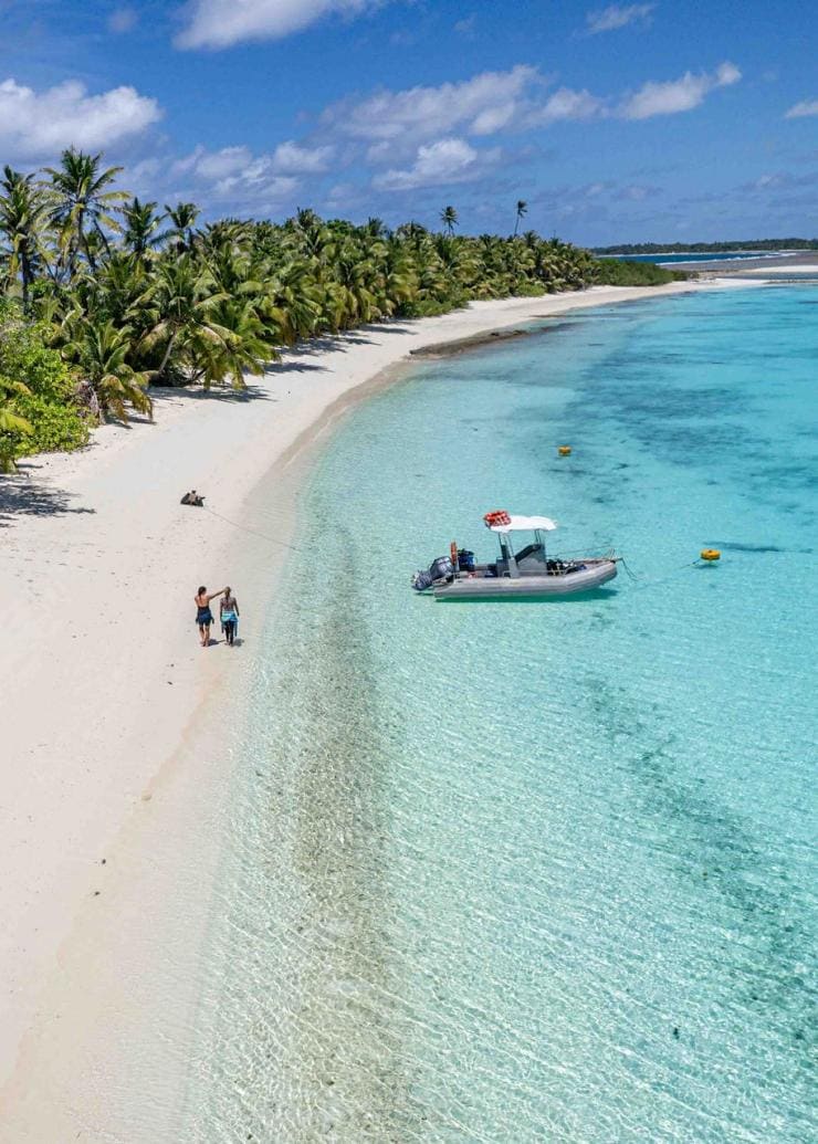 Aerial view over a white sand beach with green palm trees and clear blue water, with a small motor boat parked in the shallows and two people wandering along the sand at Cossies Beach, Direction Island, Cocos (Keeling) Islands © Strick & Fran