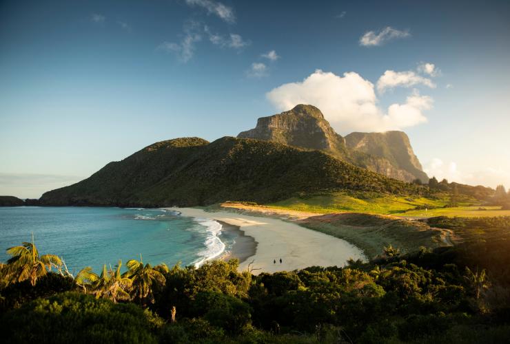 Two people walking along white sand between the blue ocean and a lush green island on Blinky Beach, Lord Howe Island, New South Wales © Tom Archer