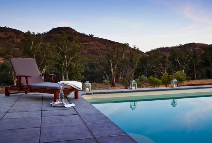 A lounge chair prepared with a towel and water glasses beside a still blue pool lined with lanterns and surrounded by greenery during dusk at Arkaba Station, Flinders Ranges, South Australia © Wild Bush Luxury