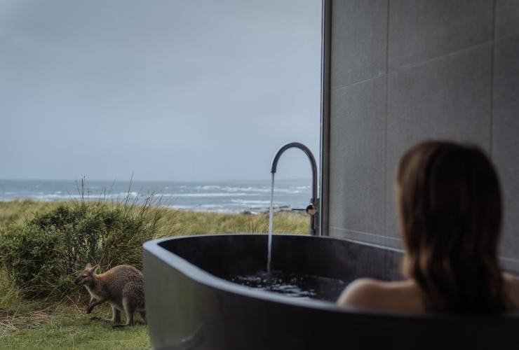 A person sitting in a large bath in front of floor-to-ceiling windows with a wallaby outside and views of the ocean beyond at Kittawa Lodge, King Island, Tasmania © Emilie Ristevski