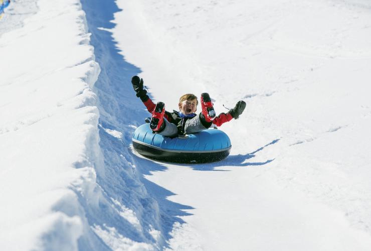 A young child smiling with their arms in the air as they ride down a snowy hill on a blue tube at Falls Creek, Victoria © Charlie Brown
