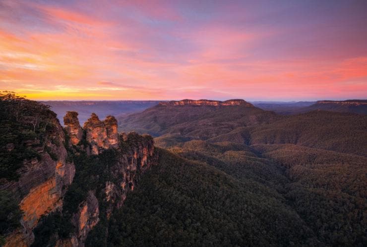 A mountain range covered in dense bushland with three towering rock formations beneath a pink and orange sky during sunset at the Three Sisters, Blue Mountains, New South Wales © Daniel Tran