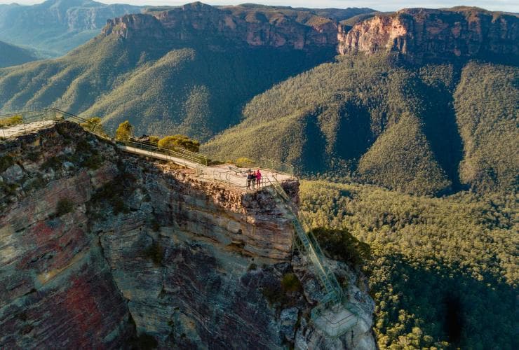 Aerial view over three people standing at the top of a sheer rocky cliff overlooking a green mountainous region from Pulpit Rock Lookout, Blue Mountains, New South Wales ©.Destination NSW/The Travel Intern