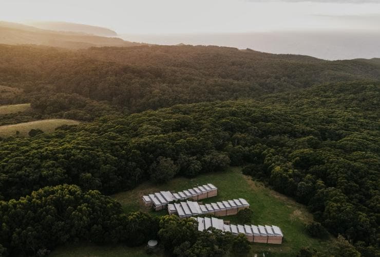 Aerial view over three rows of small buildings surrounded by greenery with the ocean visible in the distance at Alkina Lodge, Wattle Hill, Victoria © Alkina Lodge