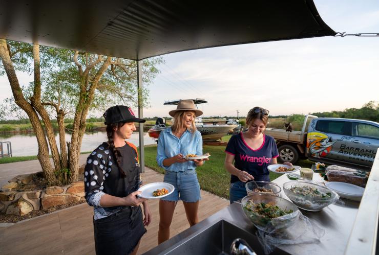 Three people helping themselves to food with a waterway and boat in the background with Barramundi Adventures Darwin, Berry Springs, Northern Territory © Tourism Australia