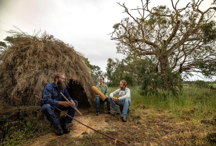 Two visitors seated around a structure made from tree branches while holding artefacts with a local Aboriginal guide during a guided tour at Budj Bim Cultural Landscape, Victoria © Visit Victoria