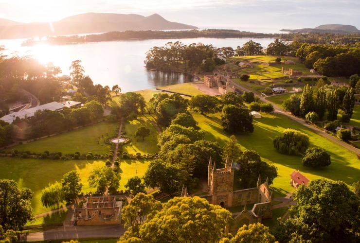 Aerial view of the sun shining over Port Arthur Historic Site, surrounded by greenery and still blue water in Port Arthur, Tasmania © Alastair Bett