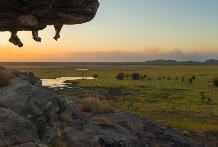 People sitting with their legs hanging over a ledge at Ubirr Aboriginal rock art site, overlooking the lush green expanse of Kakadu National Park during sunset, Northern Territory © Joe Florian