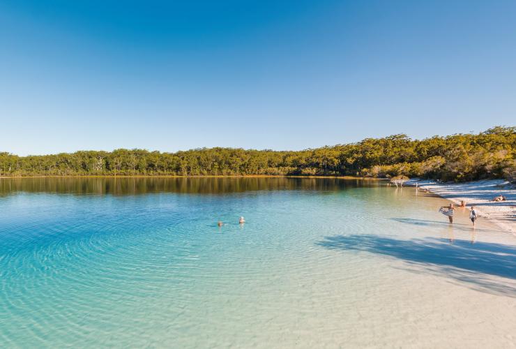 People swimming in the bright blue water of Lake Mackenzie and lazing on the white sand shores fringed by greenery on K’gari, Queensland © Tourism and Events Queensland