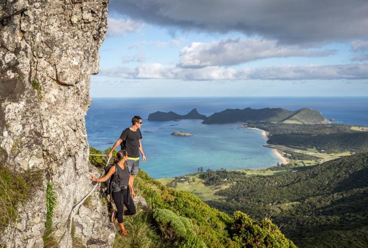 A man and woman walking on the edge of a mountain while holding onto a rope and overlooking Lord Howe Island on the Seven Peaks Walk, New South Wales © GWOA