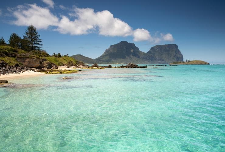 Clear blue water of the ocean in the foreground fringed by white sand with the towering green structures of Mount Lidgbird and Mount Gower in the distance on Lord Howe Island, New South Wales © Tom Archer