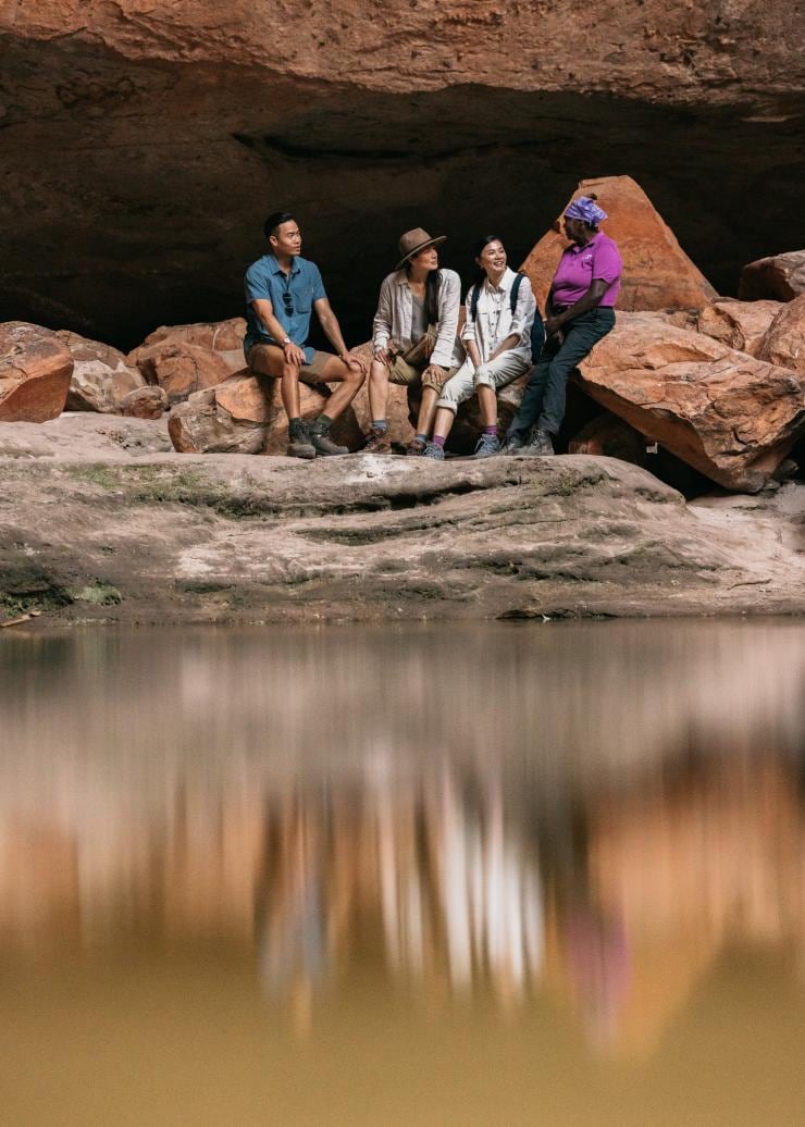 A group of people seated beside a local Aboriginal guide from Kingfisher Tours beneath a large rock formation with a waterway in front of them in Purnululu National Park, Western Australia © Tourism Western Australia