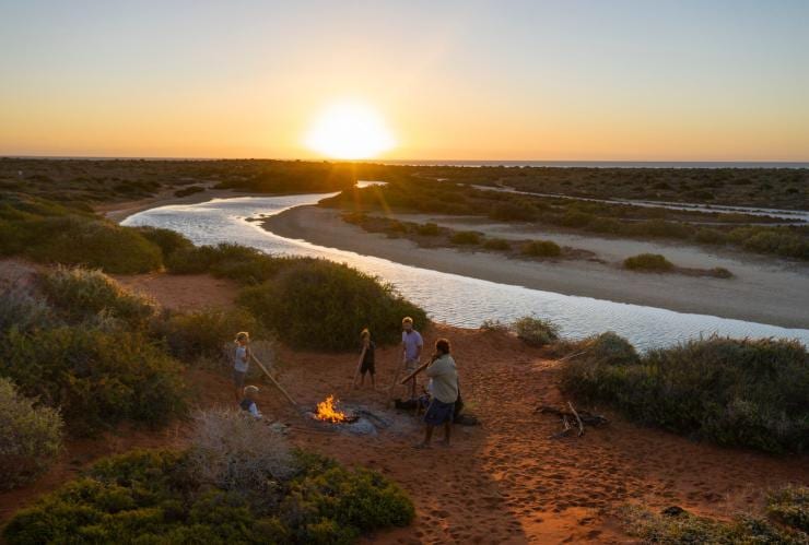 A family standing around  campfire with a local Aboriginal guide during sunset amid red sands, greenery and a waterway  with Wula Gura Nyinda Eco Adventures, Shark Bay World Heritage Area, Western Australia © Tourism Western Australia 