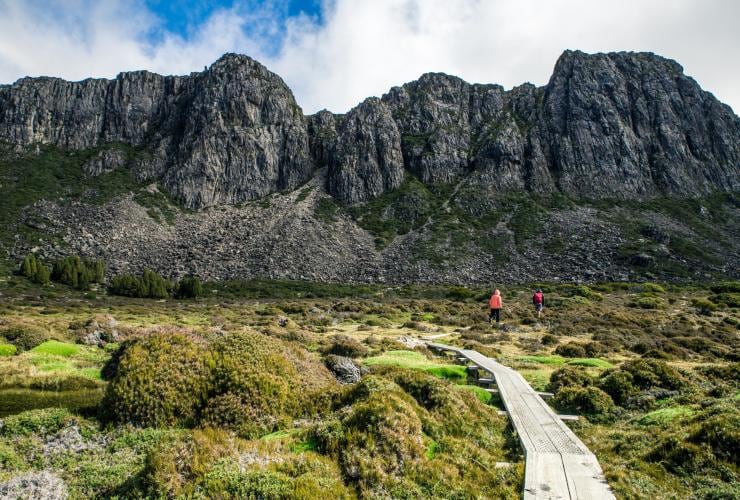 Two hikers walking along a narrow path above a sea of plants with towering rock formations in front of them in Walls of Jerusalem National Park, Tasmanian Wilderness World Heritage Area, Tasmania © O&M St John Photography