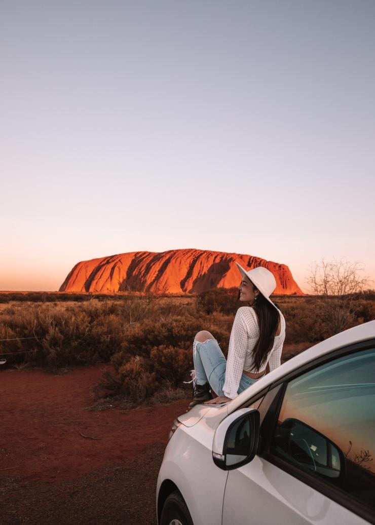 A woman sitting on the front of her car looking at Uluru as it glows red in the hues of sunset, Uluru-Kata Tjuta National Park, Northern Territory © Tourism NT/Lola Hubner