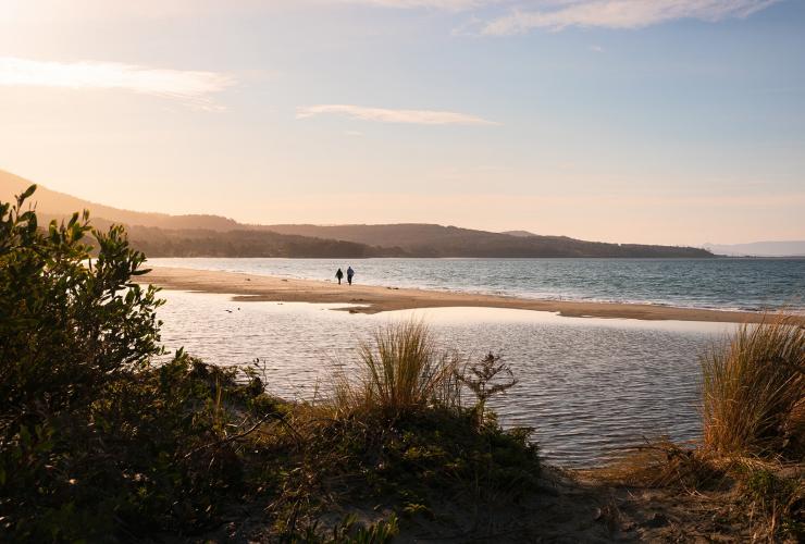 Two people walking side by side along a sand island surrounded by water with leafy green trees in the foreground during sunset at Adventure Bay, Bruny Island, Tasmania © Daniel Tran