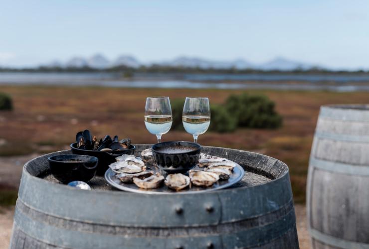 A plate of oysters on a barrel beside two glasses of white wine overlooking nature at Freycinet Marine Farm & Oyster Bay Tours, Freycinet, Tasmania © Tourism Australia