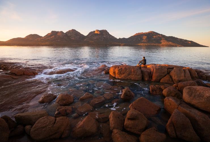 A person sitting on large rocks surrounded by the ocean with mountains in the distance at Coles Bay, Freycinet National Park, Tasmania © Daniel Tran