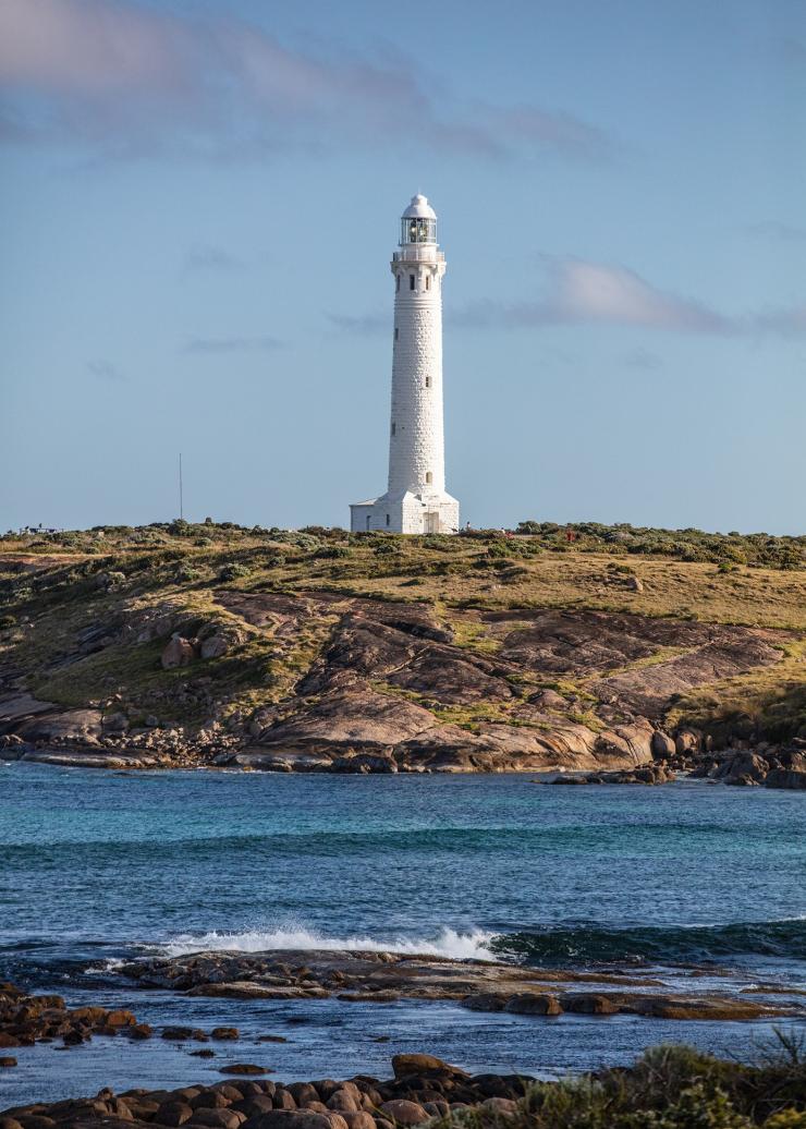 The white tower of Cape Leeuwin Lighthouse visible across a body of blue water on top of a grassy hill in Margaret River, Western Australia © Tourism Australia