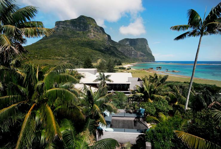 Aerial view over an outdoor seating area surrounded by palm trees with lush mountains and clear blue ocean beyond at Capella Lodge, Lord Howe Island, New South Wales © Baillie Lodges
