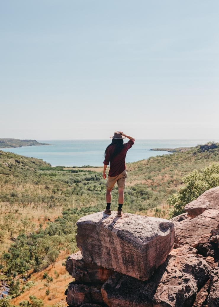 A person standing on the edge of a rocky cliff overlooking a stretch of bushland leading to the blue ocean at Faraway Bay, the Kimberley, Western Australia © Tourism Western Australia