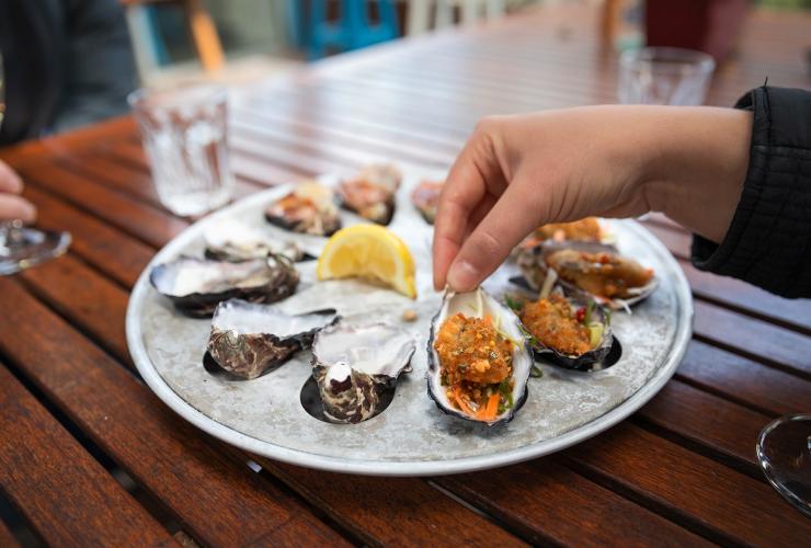Close up of a plate of oysters on ice with a person’s hand lifting a kilpatrick oyster up at Get Shucked, Bruny Island, Tasmania © Tourism Australia