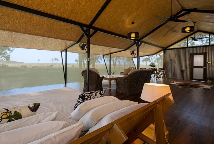 The interior of accommodation including a bed, lounges and a dining table with mesh walls giving way to views of trees and water buffalo at Bamurru Plains, Kakadu National Park, Northern Territory © Shaana McNaught