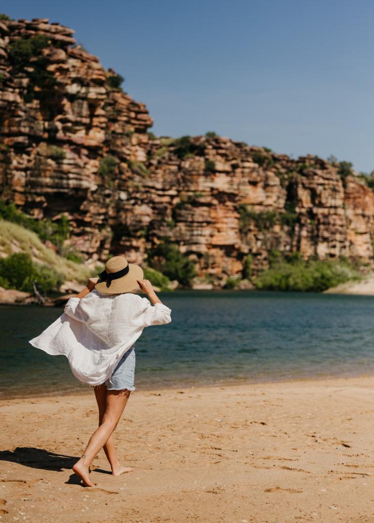 A person walking along a sandy shore with rugged cliffs across the waterway while holding their hat at Faraway Bay, the Kimberley, Western Australia © Tourism Western Australia