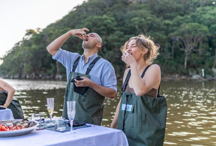 Two people wearing waders while eating oysters at a table in a river with Sydney Oyster Farm Tours, Mooney Mooney, New South Wales © Tourism Australia