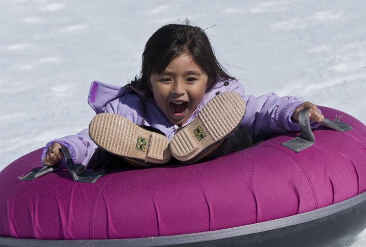 A child smiling with their mouth wide open as they slide down a snowy hill on a purple tube at Falls Creek, Victoria © Visit Victoria