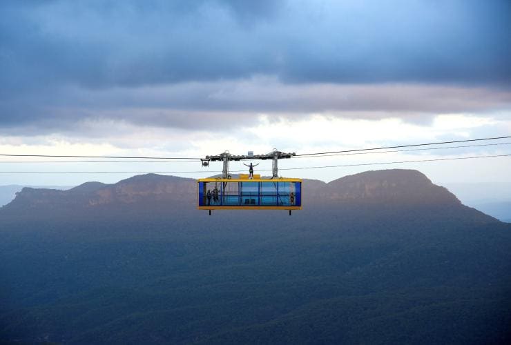 A person standing on top of a blue and yellow cable car with their arms outstretched as it travels along a wire above a hazy mountain range at Scenic World, Katoomba, New South Wales © Cam Jones Imagery
