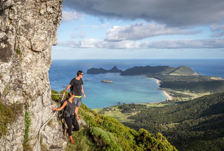 Two people standing on the edge of a cliff overlooking lush green bushland and a bright blue coast along the Seven Peaks Walk, Lord Howe Island, New South Wales © Great Walks of Australia