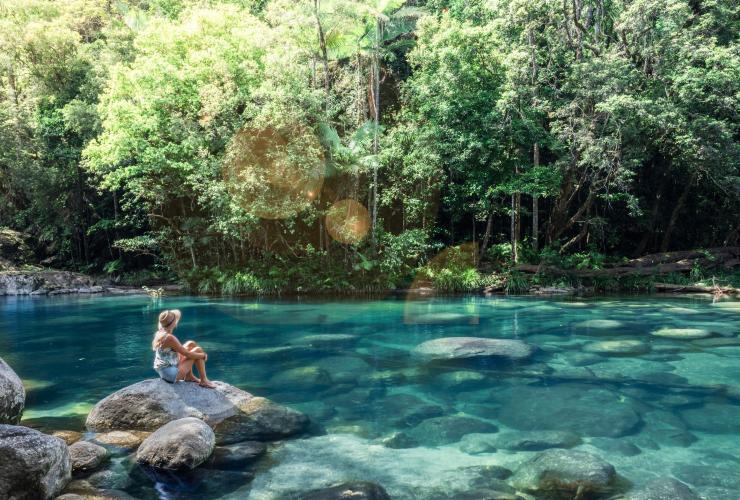 A woman perched on a large boulder in a clear blue waterhole surrounded by trees at Mossman Gorge, Mossman, Queensland © Tourism and Events Queensland