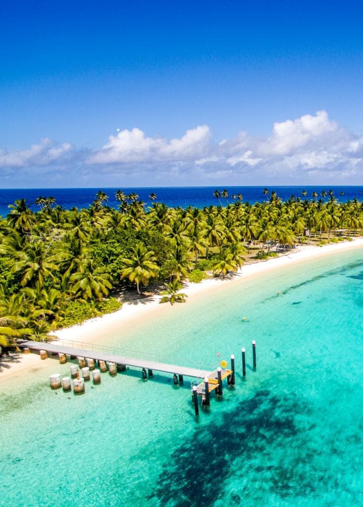 Aerial view over a white sand beach lined by green palm trees and clear, bright blue water with a pier stretching out in the foreground on Cossies Beach, Direction Island, Cocos (Keeling) Islands © Cocos (Keeling) Islands Tourism Association