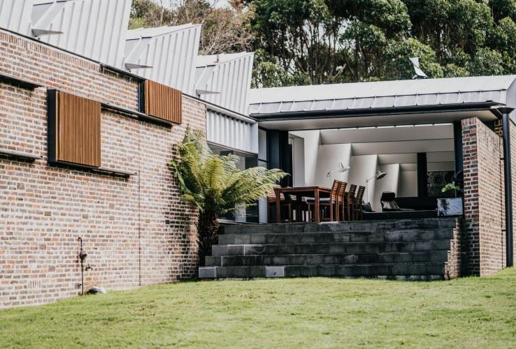 The exterior of a building with an open wall leading to a balcony with an outdoor dining table and steps down to a grassy lawn at Alkina Lodge, Wattle Hill, Victoria © Alkina Lodge