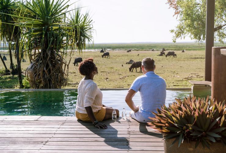 A couple seated on a deck beside a pool, with wild water buffalo grazing on the grass beyond at Bamurru Plains, Kakadu National Park, Northern Territory © Tourism NT