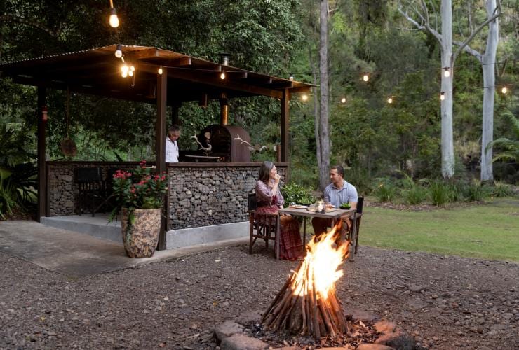 A couple dining at an outdoor table beside a campfire as a chef prepares dinner in an outdoor kitchen beside them at Nightfall Camp, Lamington National Park, Queensland © Tourism and Events Queensland