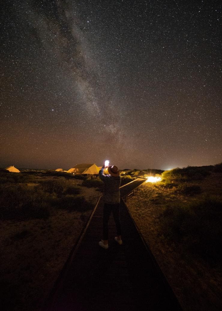 A person standing on a boardwalk at night, taking a photo of the night sky filled with stars at Sal Salis, Ningaloo Reef, Western Australia © Sal Salis Ningaloo Reef